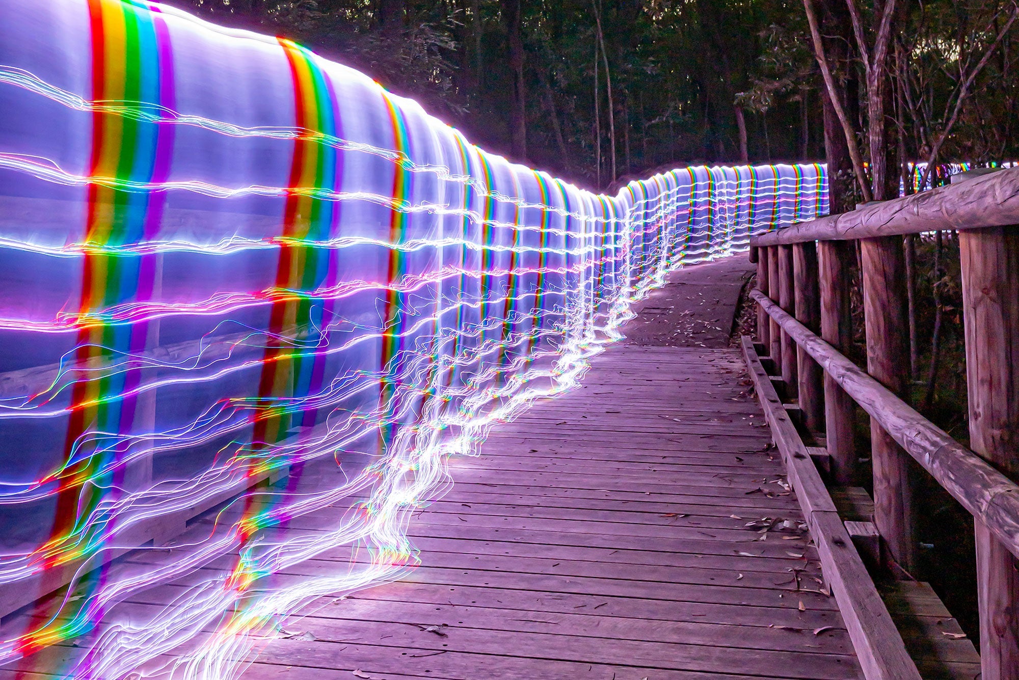 Long exposure light painting photograph of rainbow whip trails arcing along a wooden boardwalk