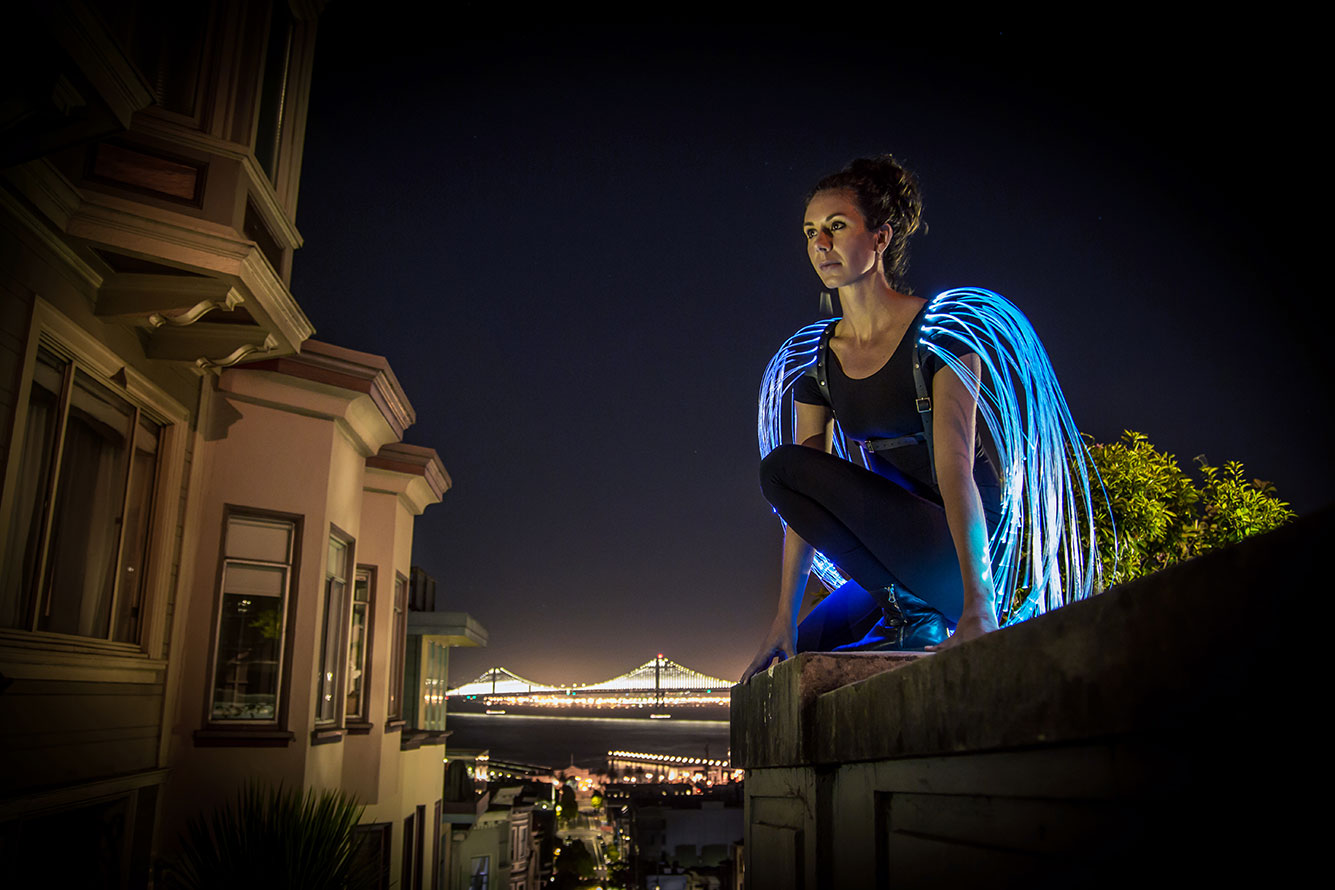 Performer wearing glowing blue fiber optic wings on a rooftop above a city skyline at night