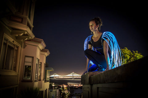 Performer wearing glowing blue fiber optic wings on a rooftop above a city skyline at night